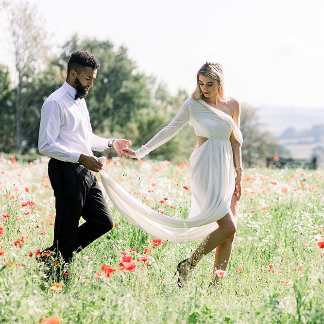 Groom in a white shirt and bow tie holding his brides wedding dress train as they walk through poppy fields 