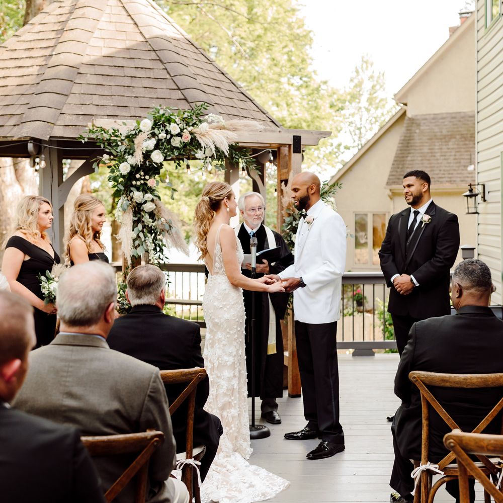 Bride in white Made With Love embroidered wedding dress holds hands with groom in white tuxedo jacket during wedding ceremony at backyard wedding