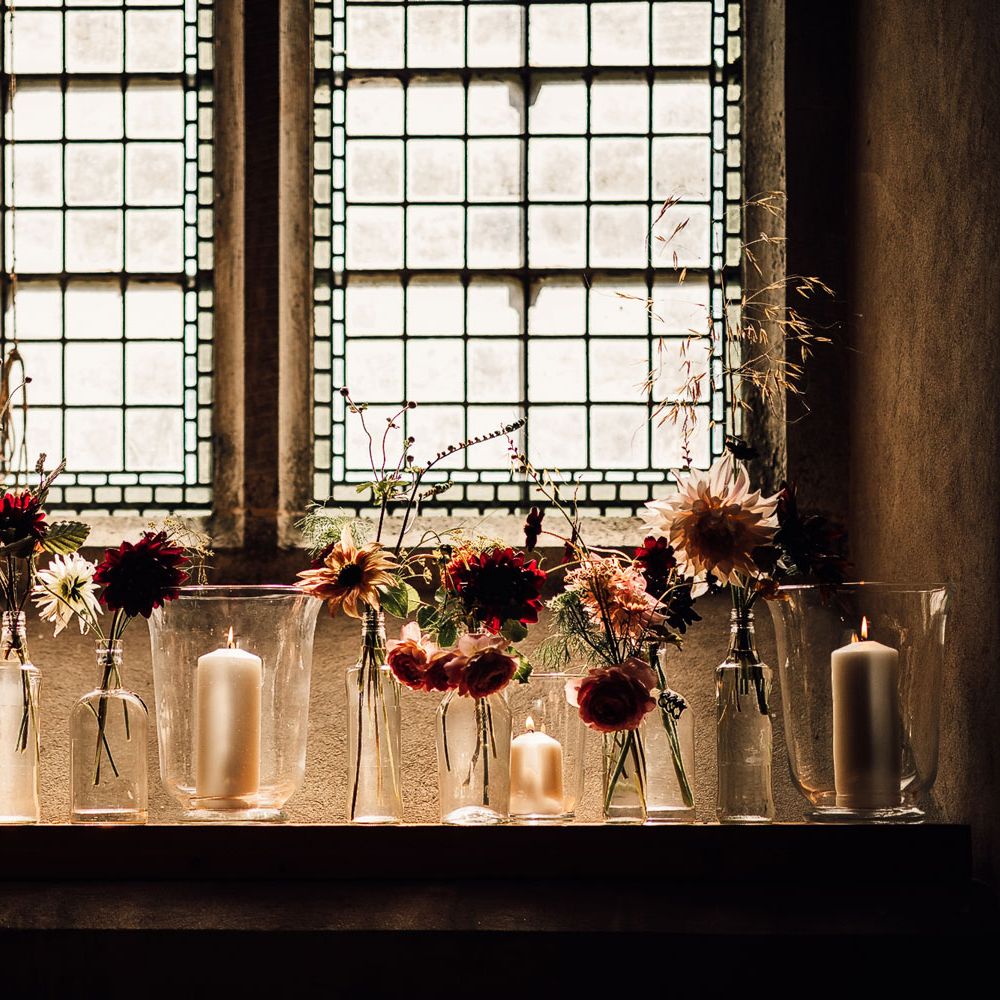 Mixed candles in glass candle holders and pink and red florals in assorted glass vases on church windowsill for Cornish wedding