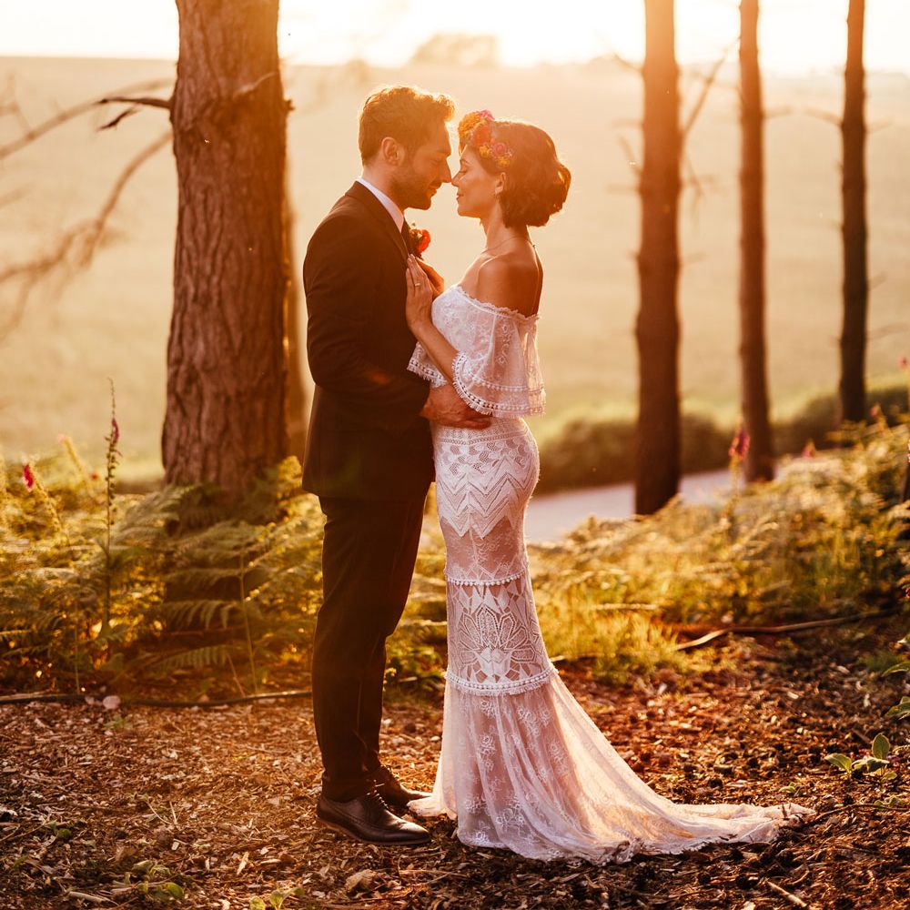 Bride in off the shoulder Grace Loves Lace wedding Dres sand flower crown stands with groom in black suit in woodland during golden hour