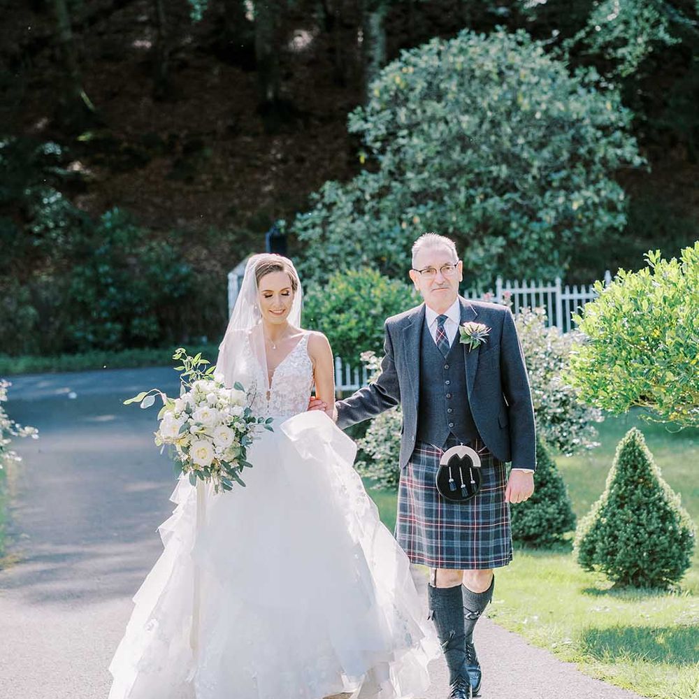 Father of the bride in a grey kilt and wool blazer escorting his daughter in a layered tulle Martina Liana wedding dress