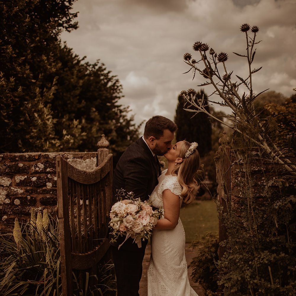 Bride in a lace Jesus Piero wedding dress and flower crown kissing her groom in the gardens at Dewsell Court, Hereford wedding venue  