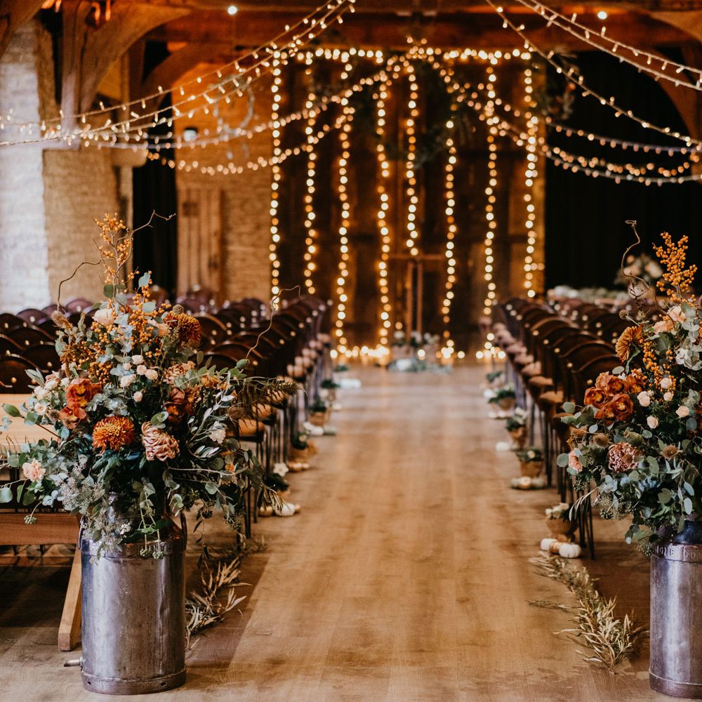 Rustic wedding aisle with milk churn floral installations, fairy light canopy and wooden chairs for rustic wedding ceremony at The Tythe Barn in Launton