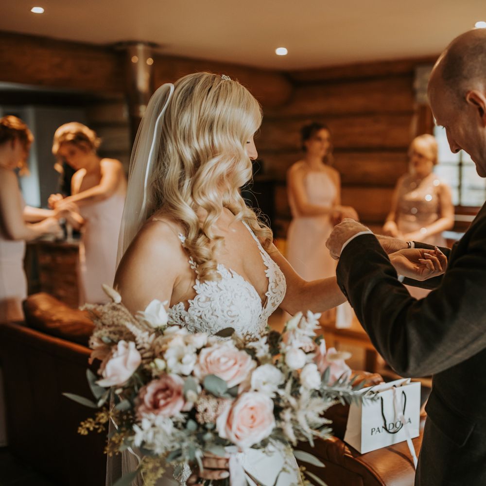 Bride with curled blonde hair, lace wedding dress and veil holding pink and white bridal bouquet has bracelet put on by father of the bride