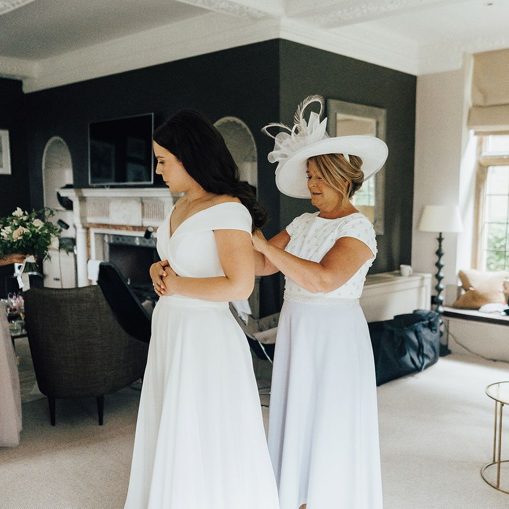 Mother of the bride in a midi dress and hat with feather doing up her daughters Suzanne Neville wedding dress on the morning of the wedding 