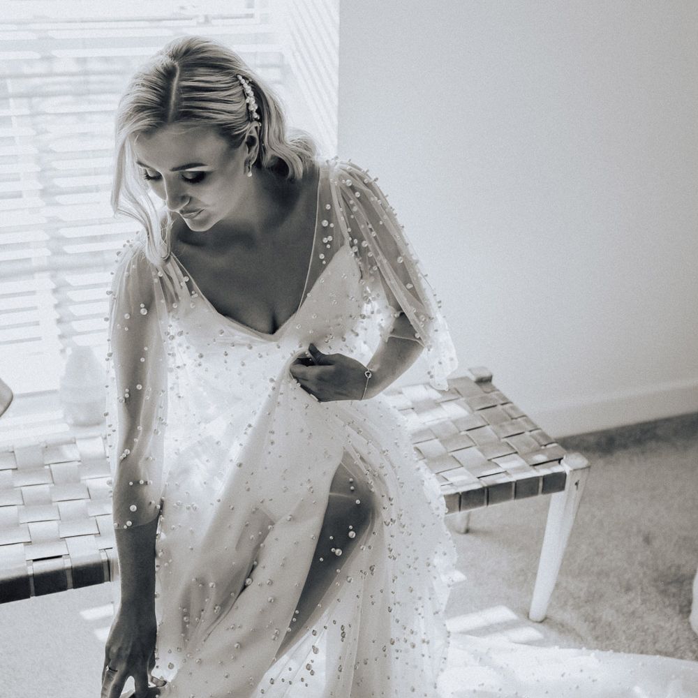 Bride in white wedding dress with mesh and pearl overlay puts on white wedding shoes with pearl strap before wedding