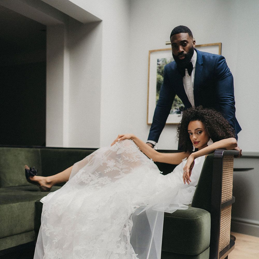 Black bride with naturally curly hair and red lipstick wearing black bow front wedding shoes sitting on a sofa in her embroidered lace wedding dress 