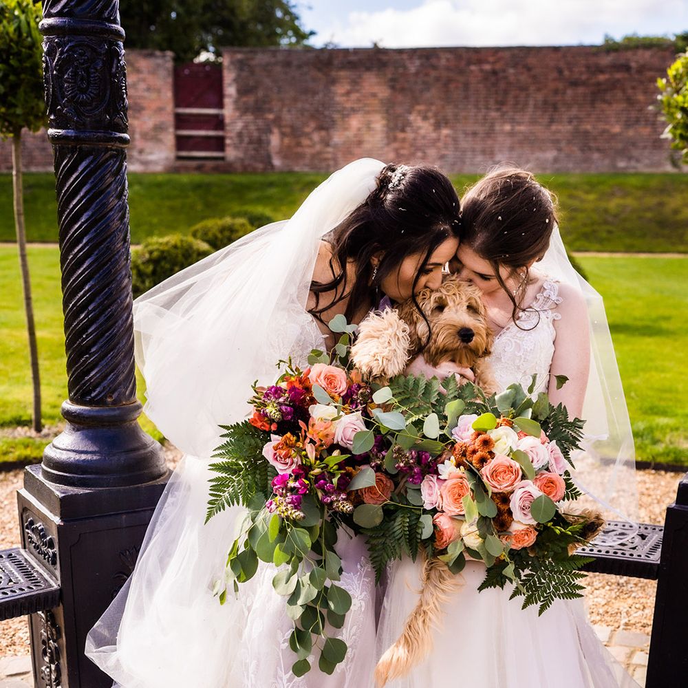 Brides in white wedding dresses holding matching wedding bouquets kiss their pet dog who was ring bearer during their ceremony