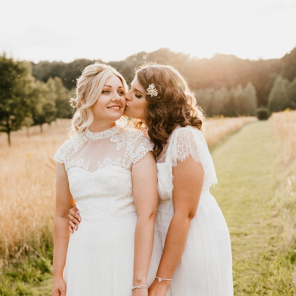 Brunette bride kisses blonde bride on the cheek after their wedding as they stand in a field together