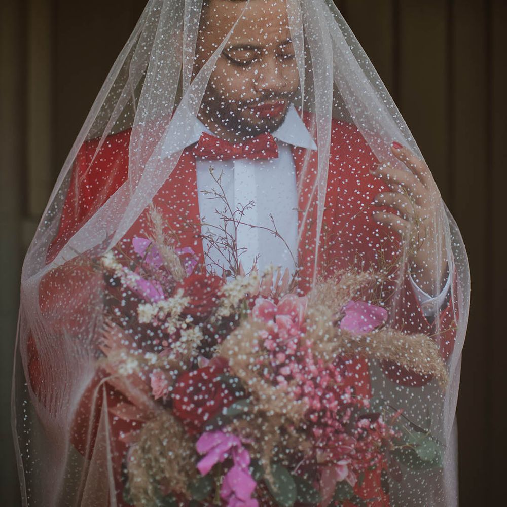 Groom in a red suit and bow tie wearing winged eyeliner and an embroidered veil over his face 