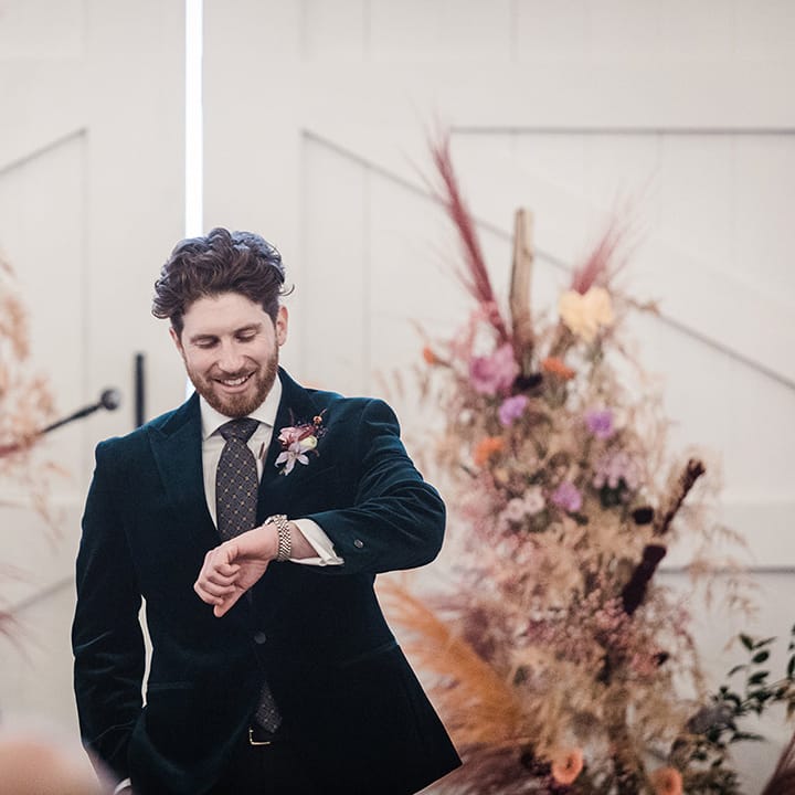 Groom in velvet suit checks his watch for the time as he stands in front of colourful dried flower columns on wedding day 