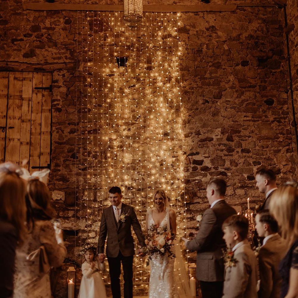 Bride and groom stand in front of fairy light backdrop at Eden Barn with the groom holding hands with their daughter in white dress and flower crown