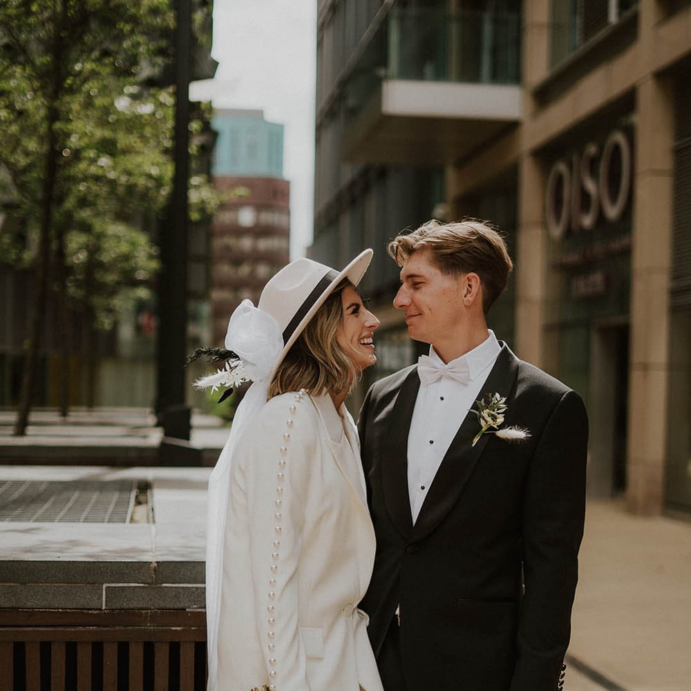 Bride and groom smile and look into each other's eyes with bride in Nadine Merabi pearl suit and groom in black tie 