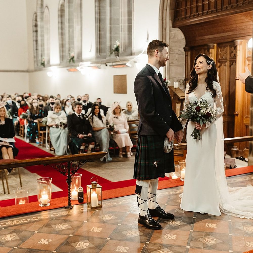 Bride wears long sleeved lace gown and groom wears tartan kilt as they stand at the altar in church during wedding ceremony 