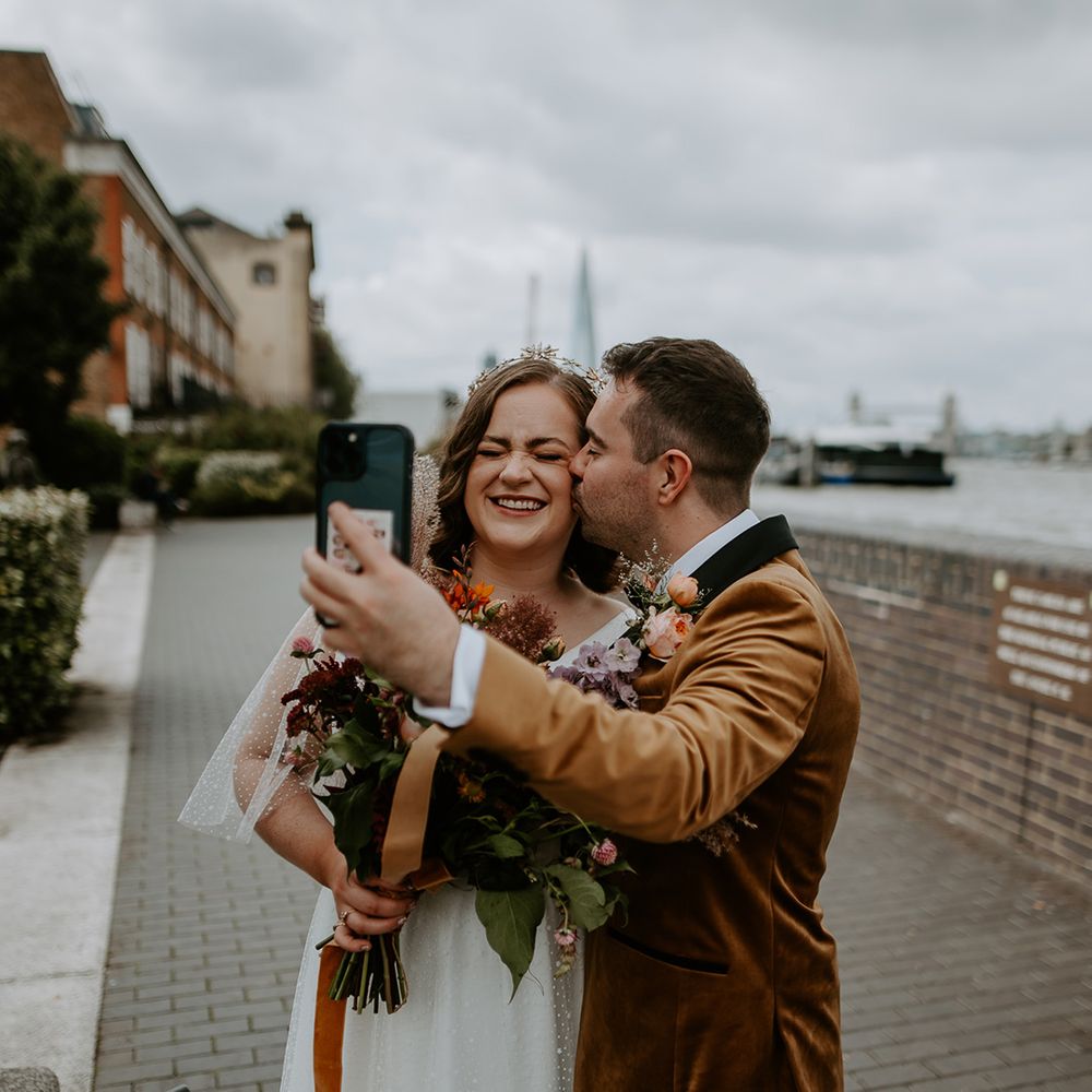 Groom kissed Bride on cheek whilst taking a selfie with Burnt orange themed bouquet and suit