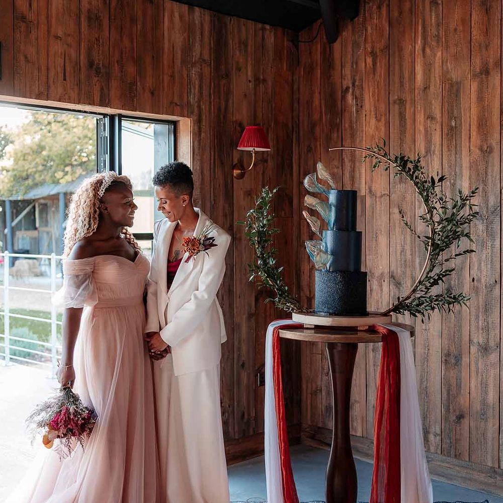 String trio playing the violin and the cello by three tier wedding cake in a dark blue buttercream with a velvet base tier and gold rice paper sails and semi circle arch stand and brides in complimenting boho bridal outfits 