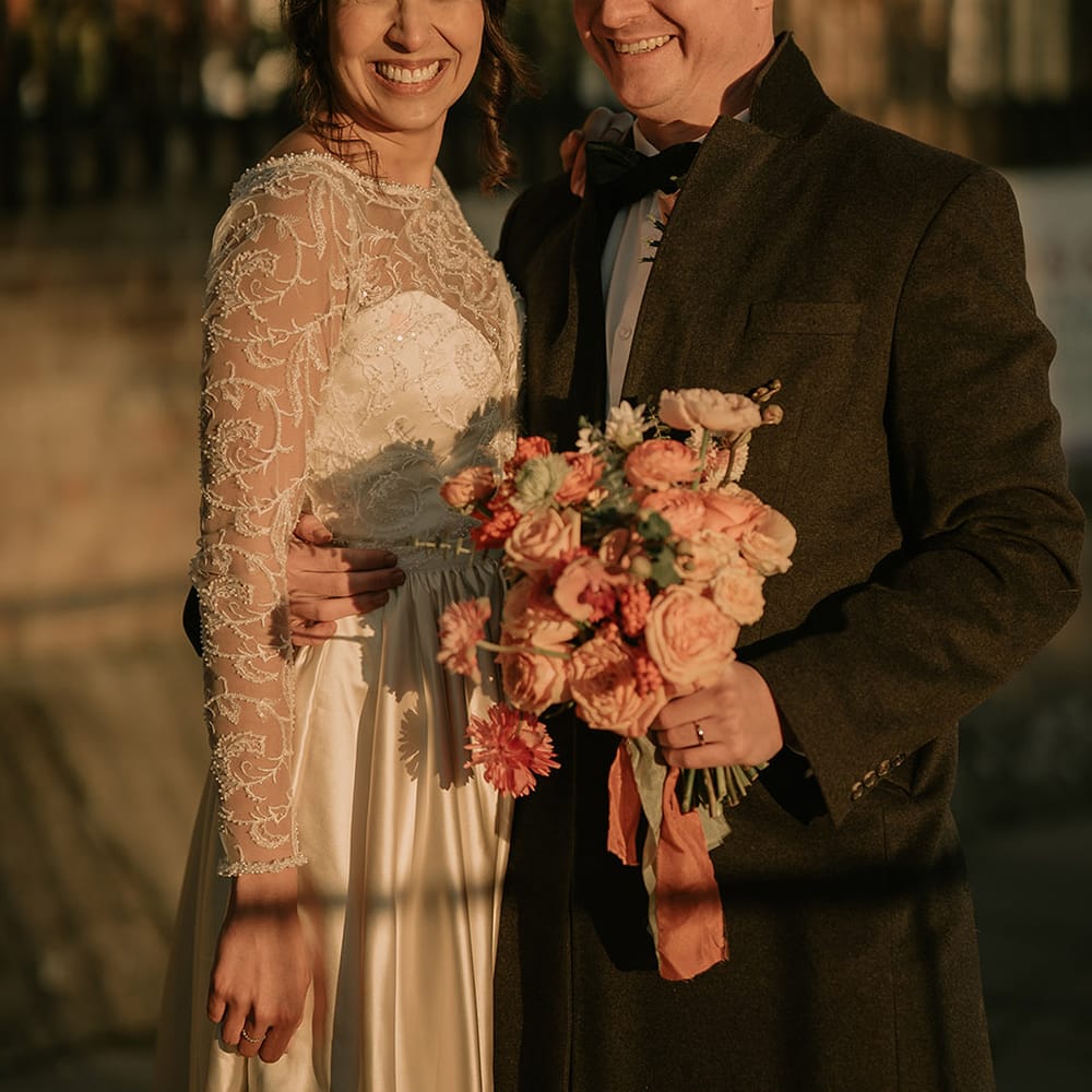 Bride in gold, pearl and emerald bridal crown wearing princess wedding dress with long lace sleeves and groom in black tux holding bouquet with light pink poppies, orange garden roses, eucalyptus and dried flowers