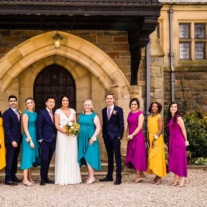 Bride & groom stand with their wedding party of grooms in blue suits beside bridesmaids in yellow, purple and teal mismatched bridesmaid dresses