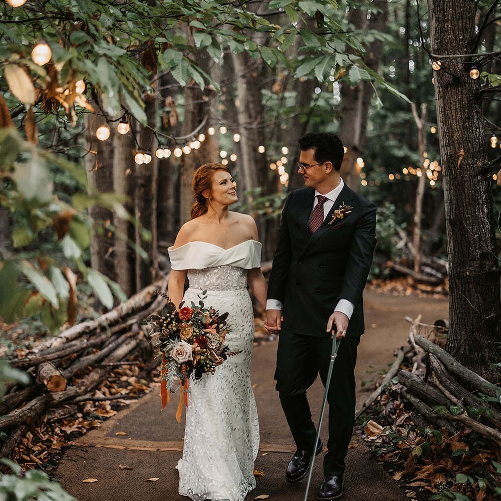 Bride & groom walk through woodland complete with festoon lighting at The Dreys wedding venue