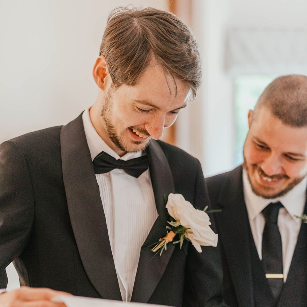 The groom in a black tuxedo with white rose buttonhole reading surprise wedding newspaper from the bride 