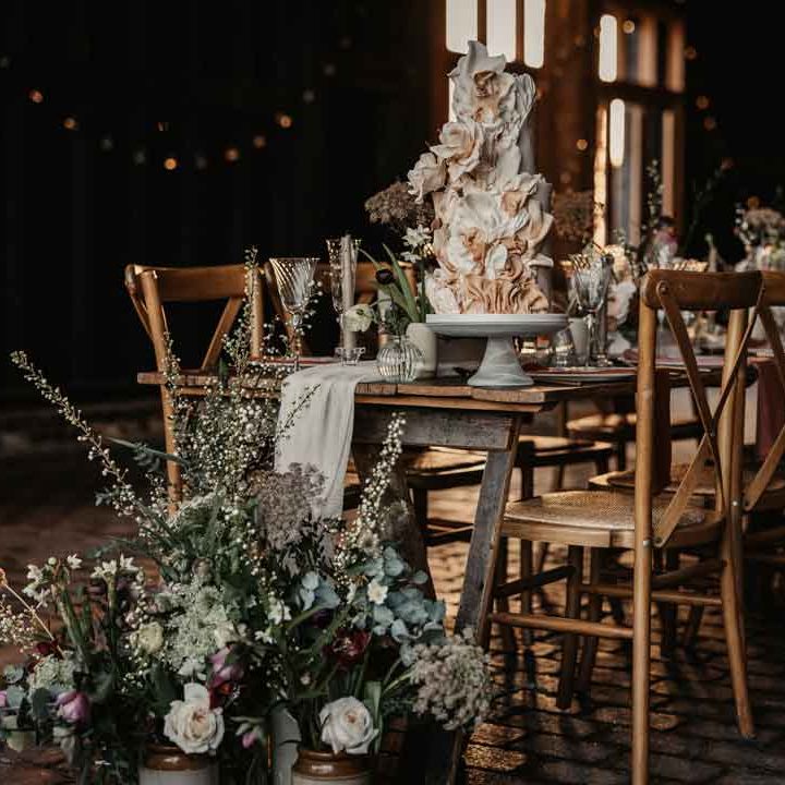 Modern rustic luxe wedding tablescape with white fabric table runner, large foliage, baby's breath, garden rose and eucalyptus floral arrangements, a large artistic layered wedding cake, tapered candles and ceramic vases 