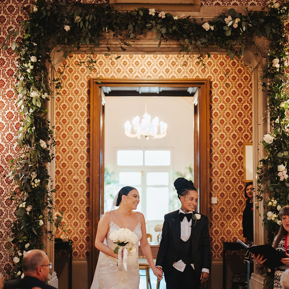 Bride in lace sleeveless v neck Enzoani wedding dress with tulle finish and church length veil and bride in black snakeskin print tuxedo with black bowtie and white rose boutonniere doing wedding exit through Grittleton House with white rose and foliage flower arch around the door 