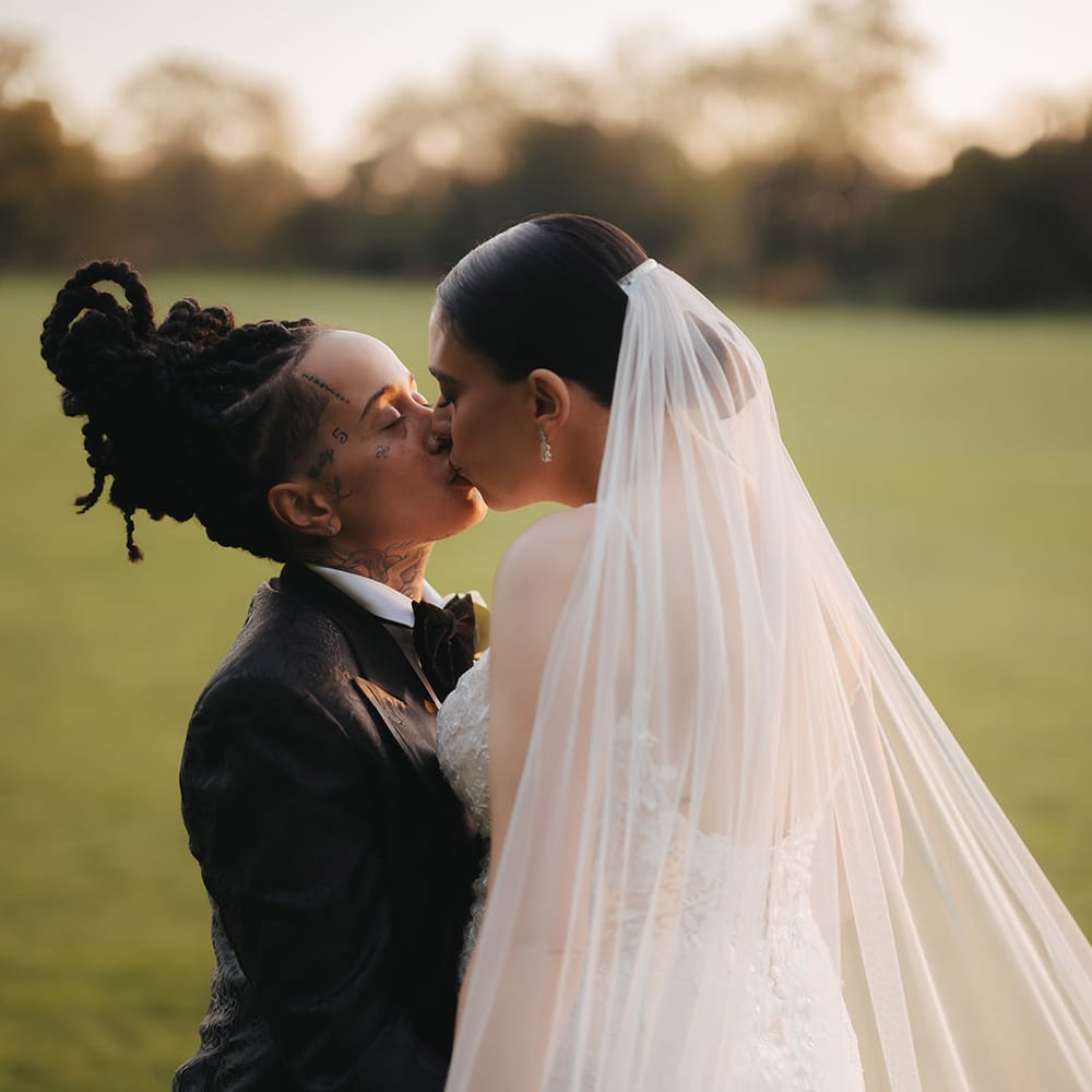Bride in black snakeskin print tuxedo with black bowtie kissing bride in lace sleeveless v neck Enzoani wedding dress with tulle finish and church length veil on the grounds of Grittleton House wedding venue 