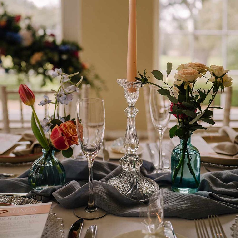 Clear candlestick holder with pastel tapered candle, bright blue glass bottles with dried flowers and champagne glasses on classic white wedding tablescape 