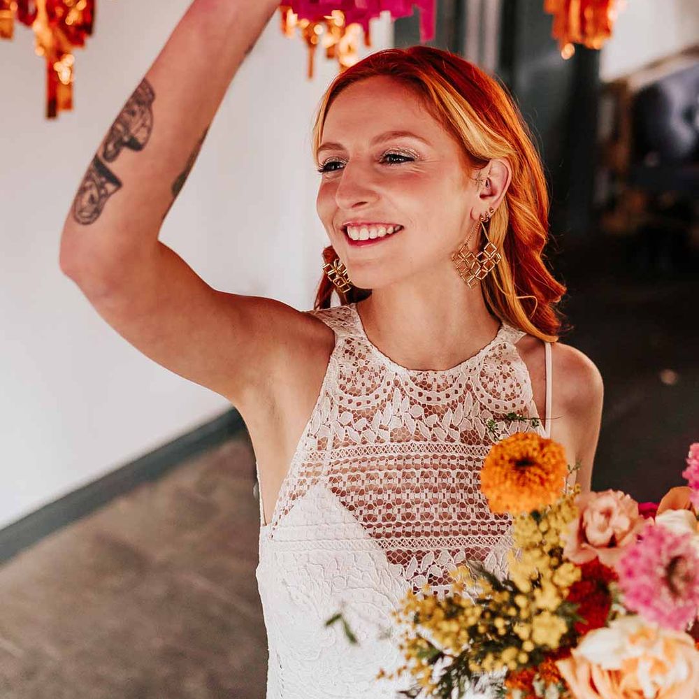 Bride in 70s inspired boatneck short lace wedding dress holding mixed flower bridal bouquet touching pink retro wedding streamers 