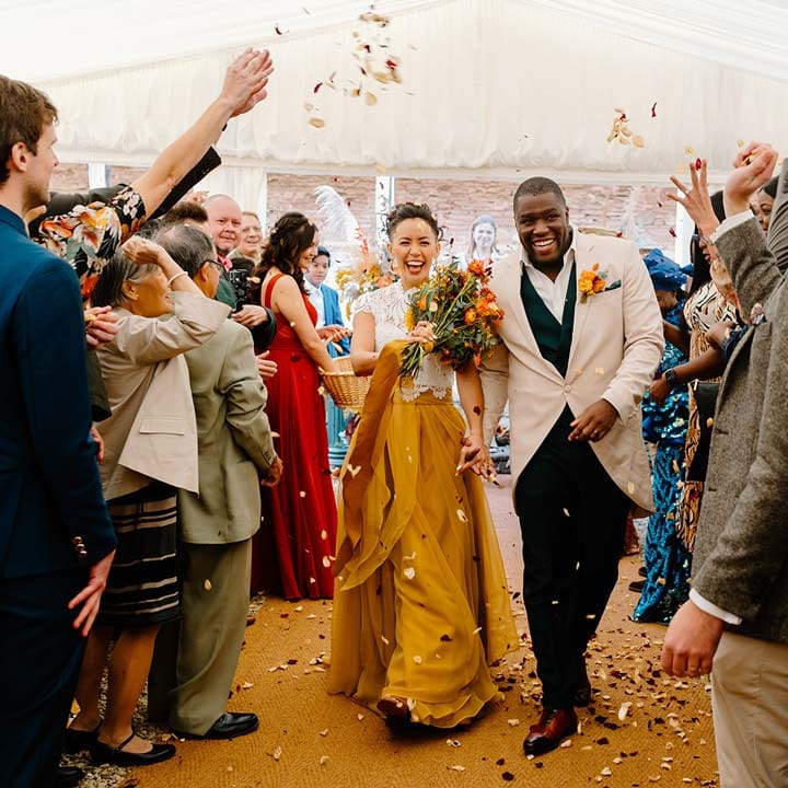 Bride in white mesh lace bridal crop top and yellow wedding skirt walking with groom in cream morning suit with forest green grooms waistcoat and trousers, forest green pocket square and orange boutonniere at Broadfield Court wedding doing colourful dried petal confetti exit