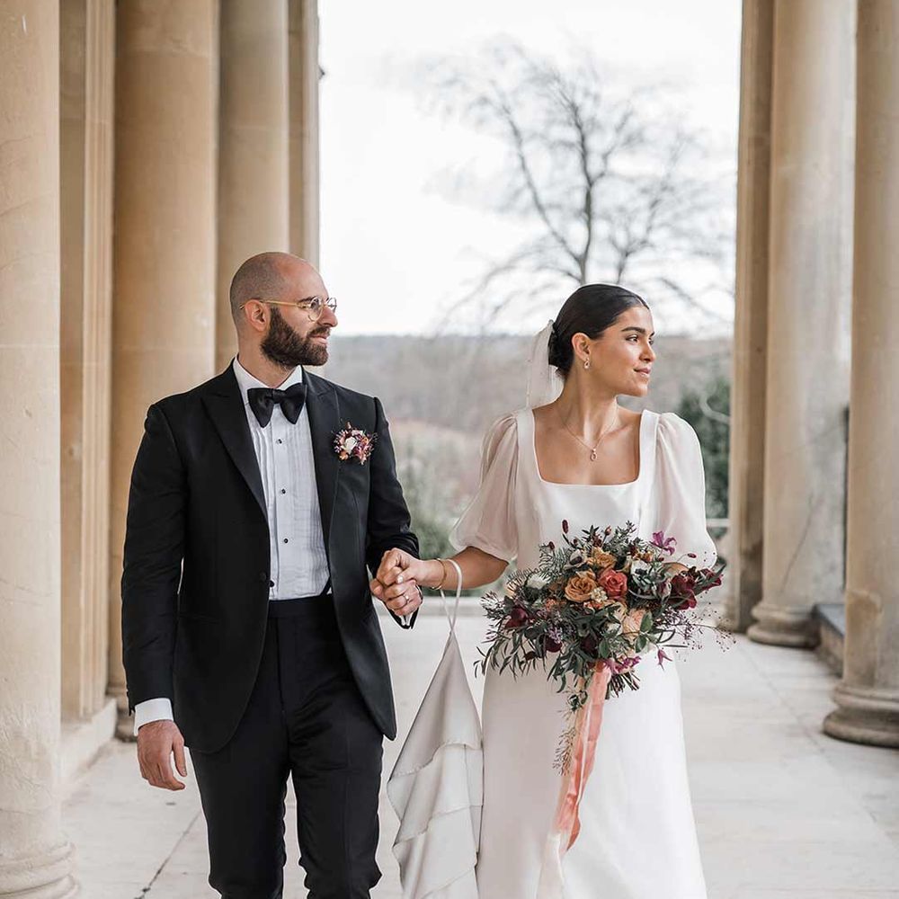 Bride in short sheer puff sleeve wedding dress with square neck and wrist loop holding dried wildflower bouquet walking with groom in classic black tuxedo with bowtie and wildflower boutonniere walking hand in hand at Buxted Park wedding venue