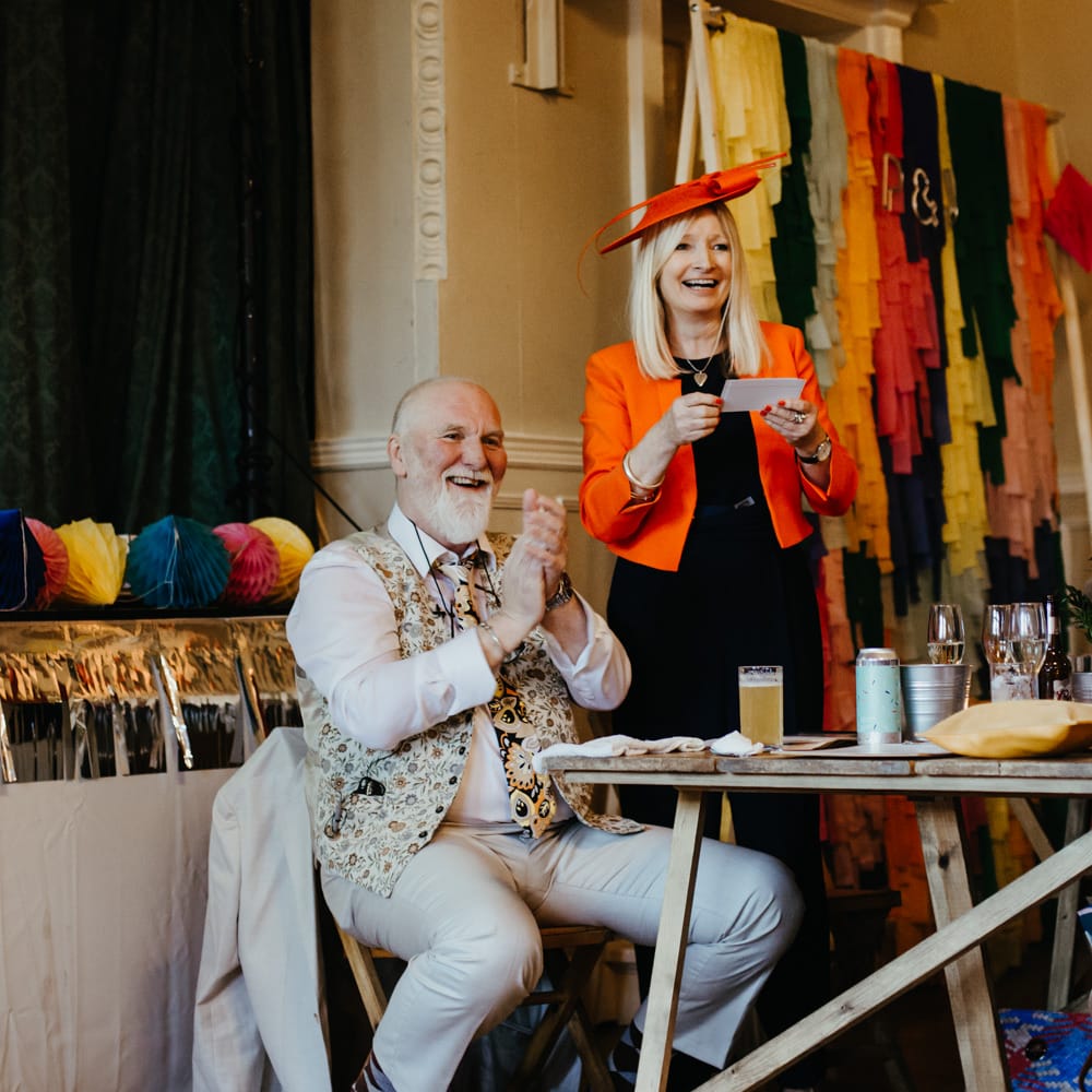 Mother of the bride in a black dress, orange jacket and hat reading a speech 