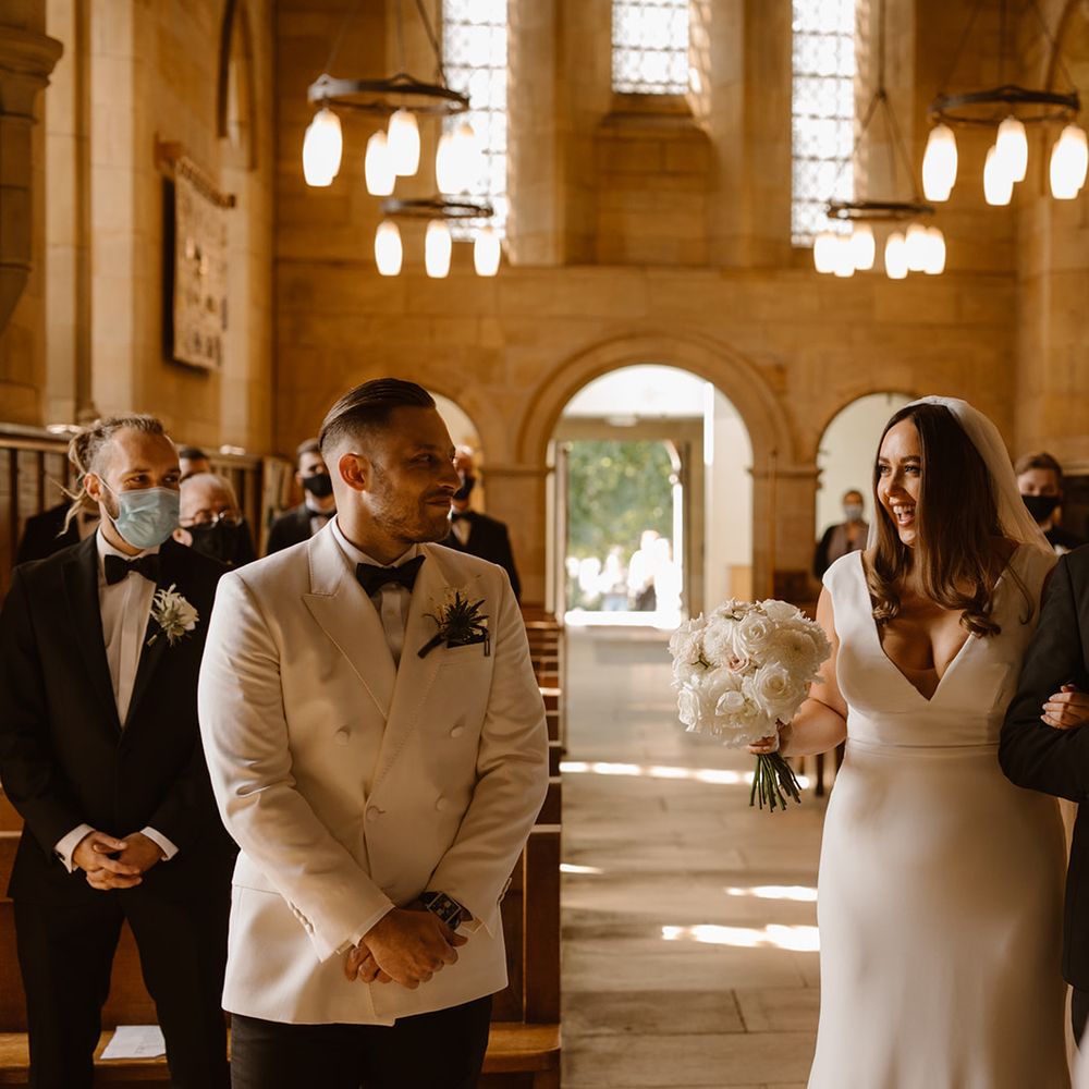 Smiling bride in Made With Love wedding dress stands arm in arm with father in black tuxedo at the church altar next to groom in white double breasted tuxedo
