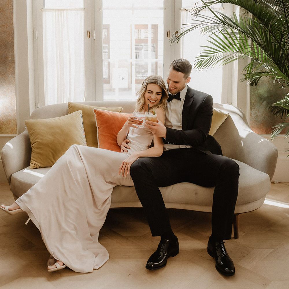 Bride and groom toasting with pink and peach coloured cocktails on a luxe sofa