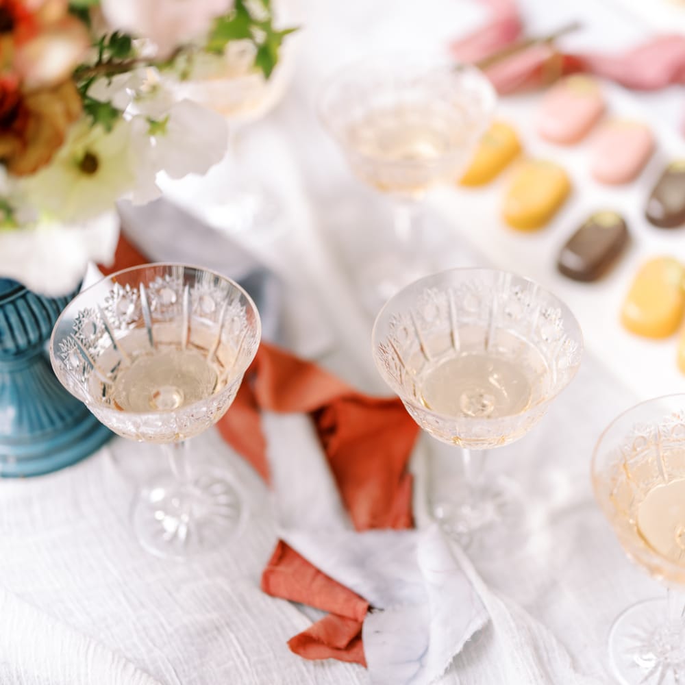 Three glasses of champagne sit on white tablecloth next to pastel coloured biscuits 