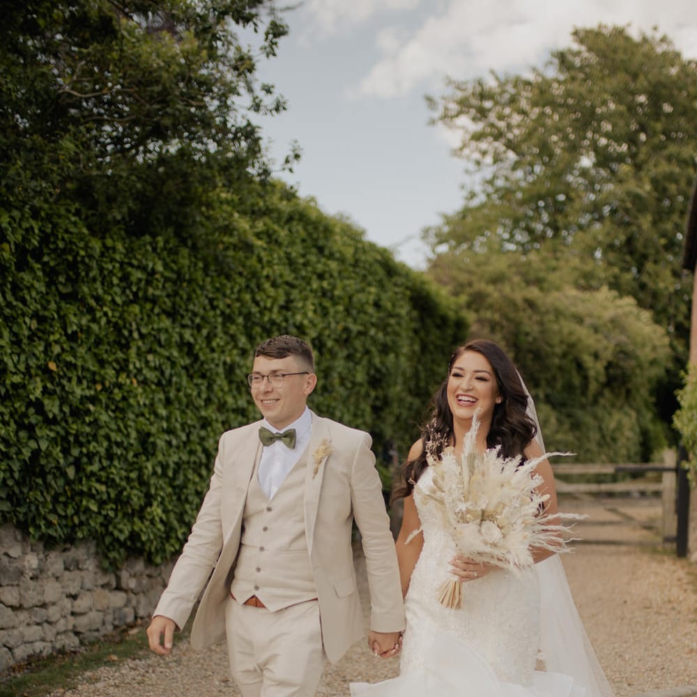 Groom in a three piece beige suit with sage green bow tie holding hands with his bride in a fishtail wedding dress