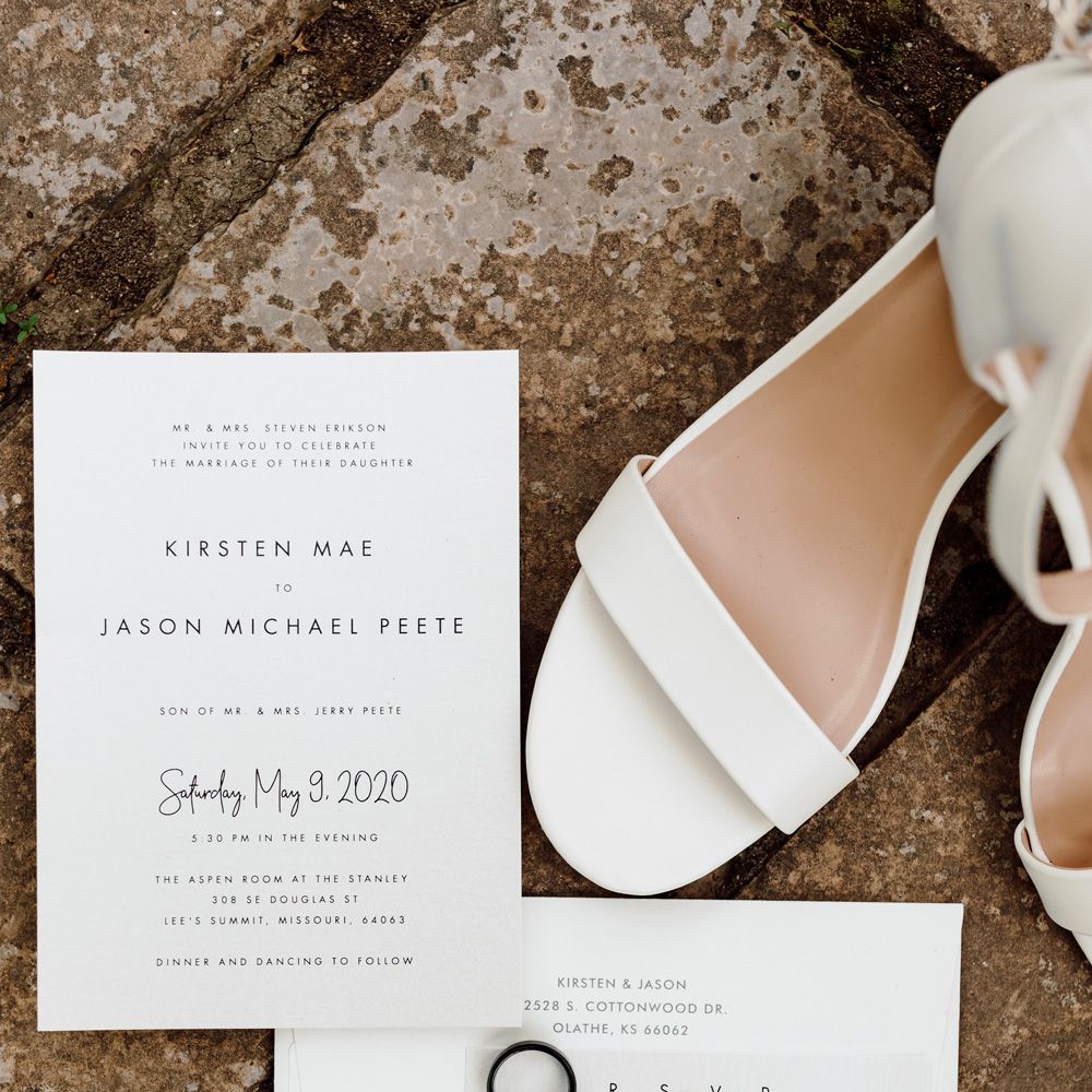 White heeled sandals with strap, white wedding invitations and wedding rings on stone floor at backyard wedding