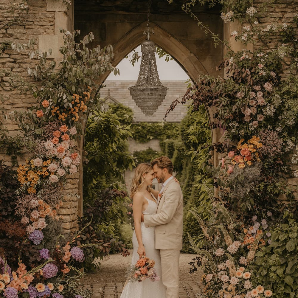 Bride and groom portrait under the castle arch with chandelier at Euridge Manor surrounded by pastel coral, pink, orange and peach wedding flowers 