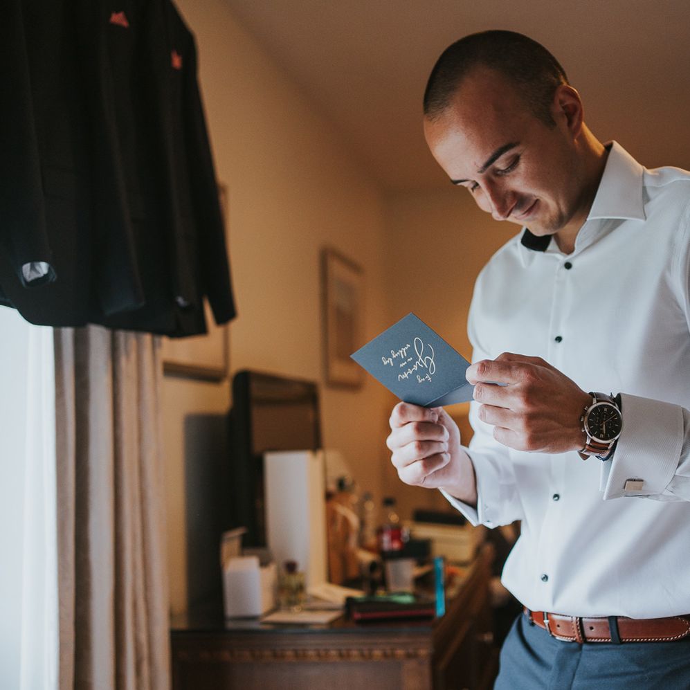 Groom in white shirt and blue suit trousers with brown belt reads message before Tythe Barn wedding with barn wedding flowers