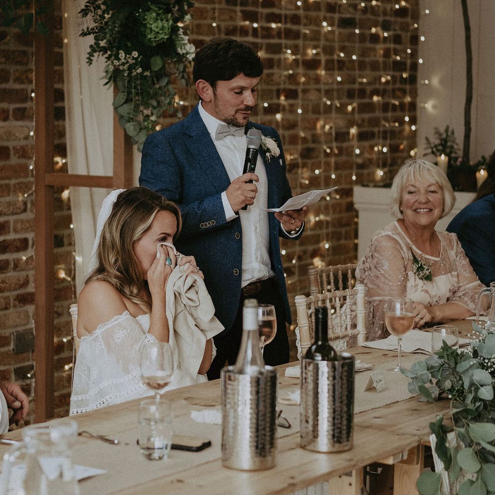 Bride in Grace Loves Lace bardot wedding dress and veil wipes eyes with napkin as groom in blue suit and bow tie makes speech at Isle of Wight wedding with macrame wedding decor