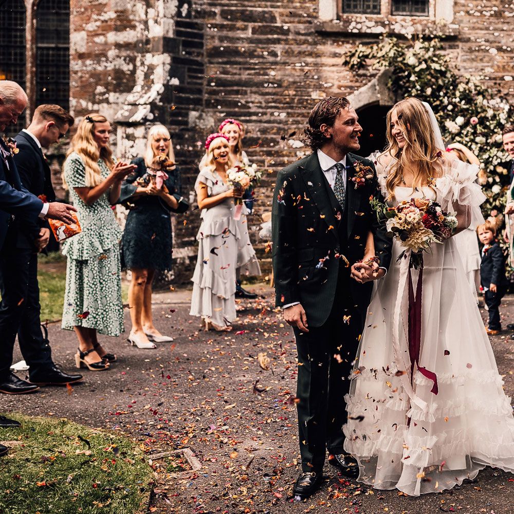 Bride in white Halfpenny London Mayfair dress holding mixed bridal bouquet with burgundy ribbon smiles holding hands with groom in three piece suitas they leave church and guests throw confetti at Cornish wedding