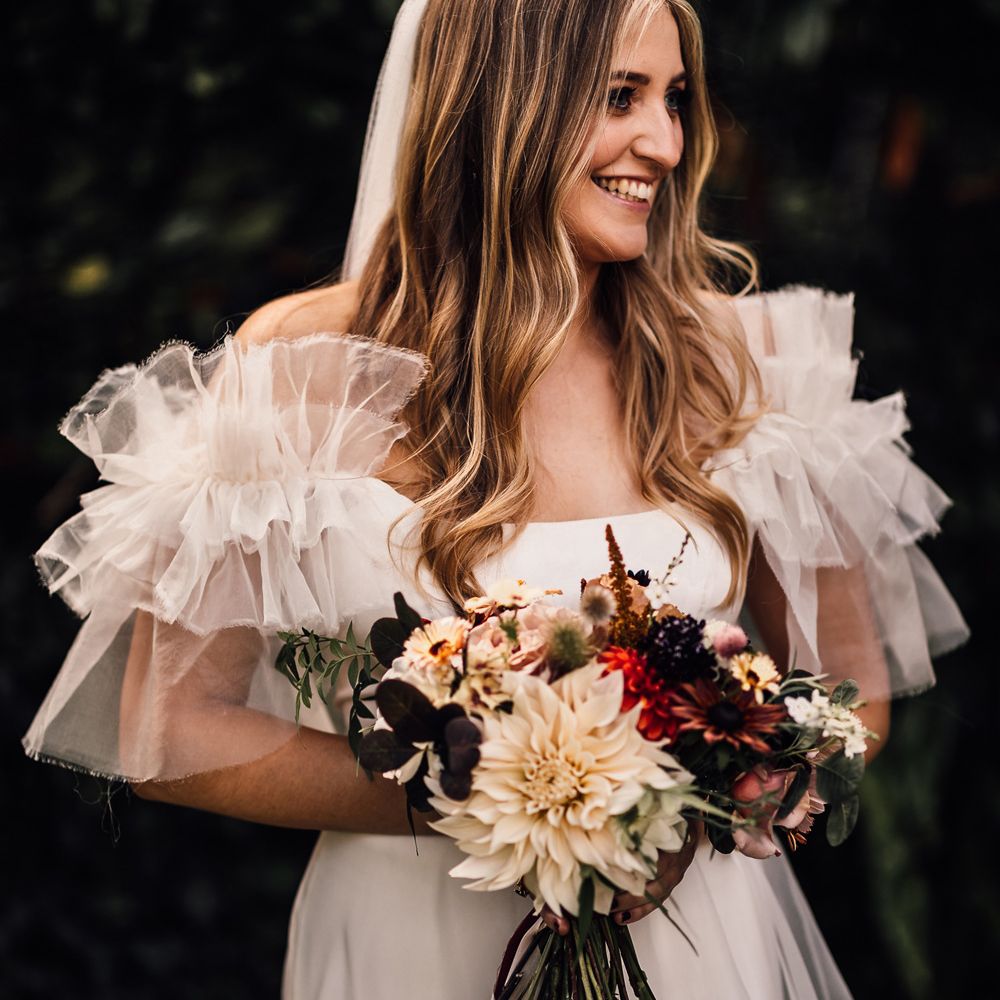 Bride in white Halfpenny London Mayfair dress with ruffle sleeves and veil stands holdingg mixed bridal bouquet with burgundy ribbon at garden wedding reception in Cornwall