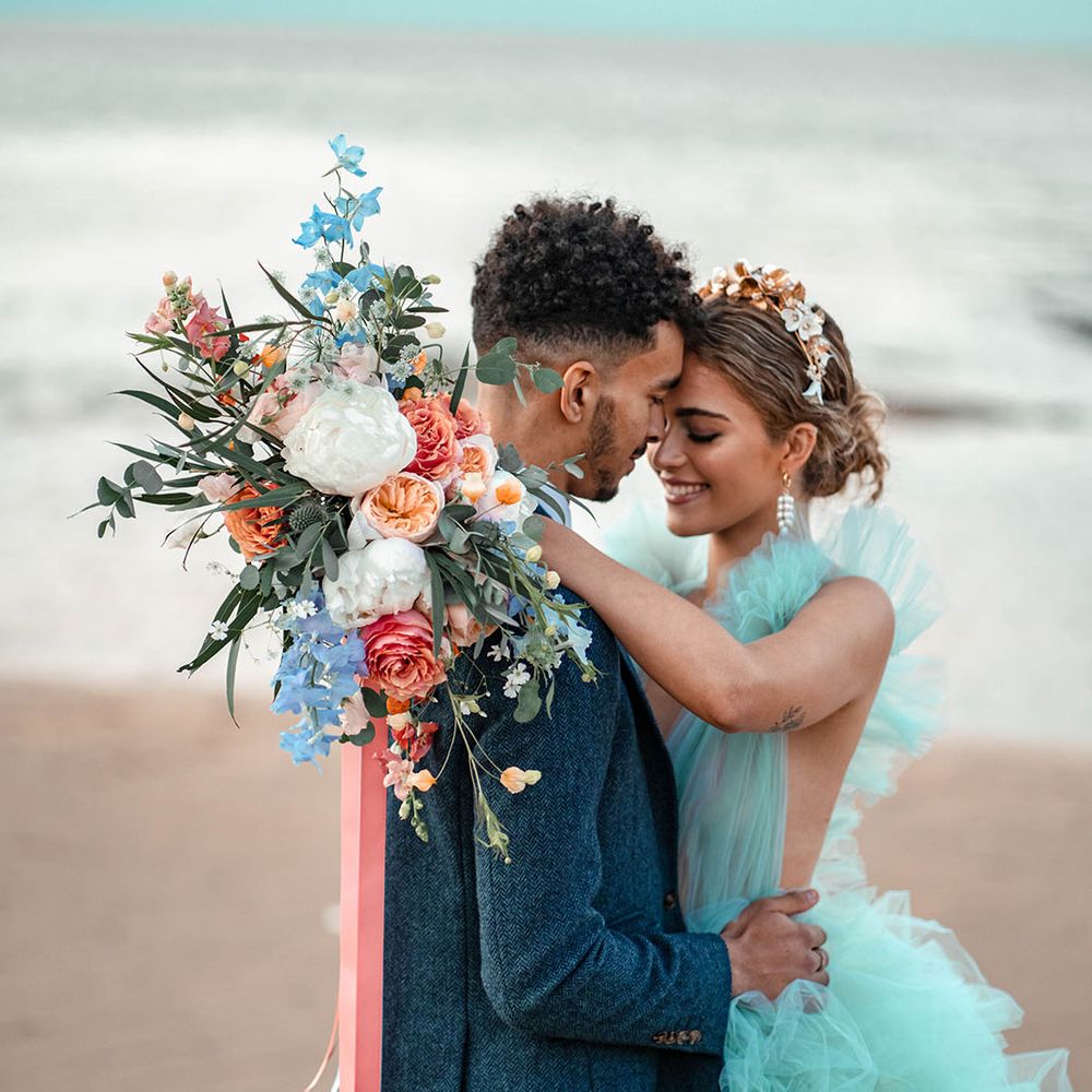Intimate coastal wedding with bride in a mint green wedding dress holding a pastel flower wedding bouquet tied with ribbon over her grooms shoulder in a blue wool suit 
