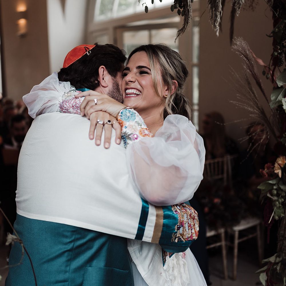 Bride in mesh sleeve wedding dress with embroidered floral cuffs hugs groom in red kippah, tallit and green suit during wedding ceremony