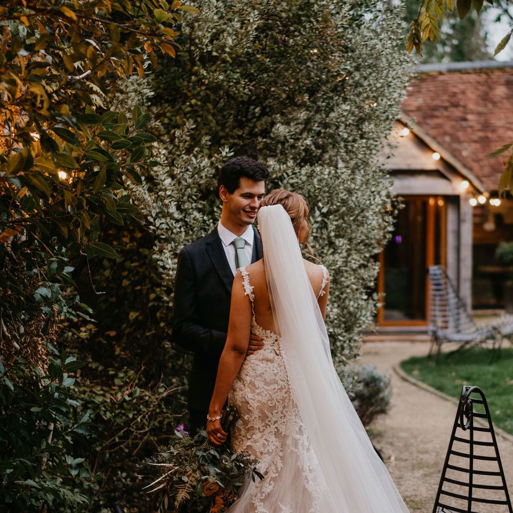 Bride in open back lace and tulle wedding dress with train and veil stands with groom in dark suit and sage green tie in the grounds of The Tythe Barn in Launton
