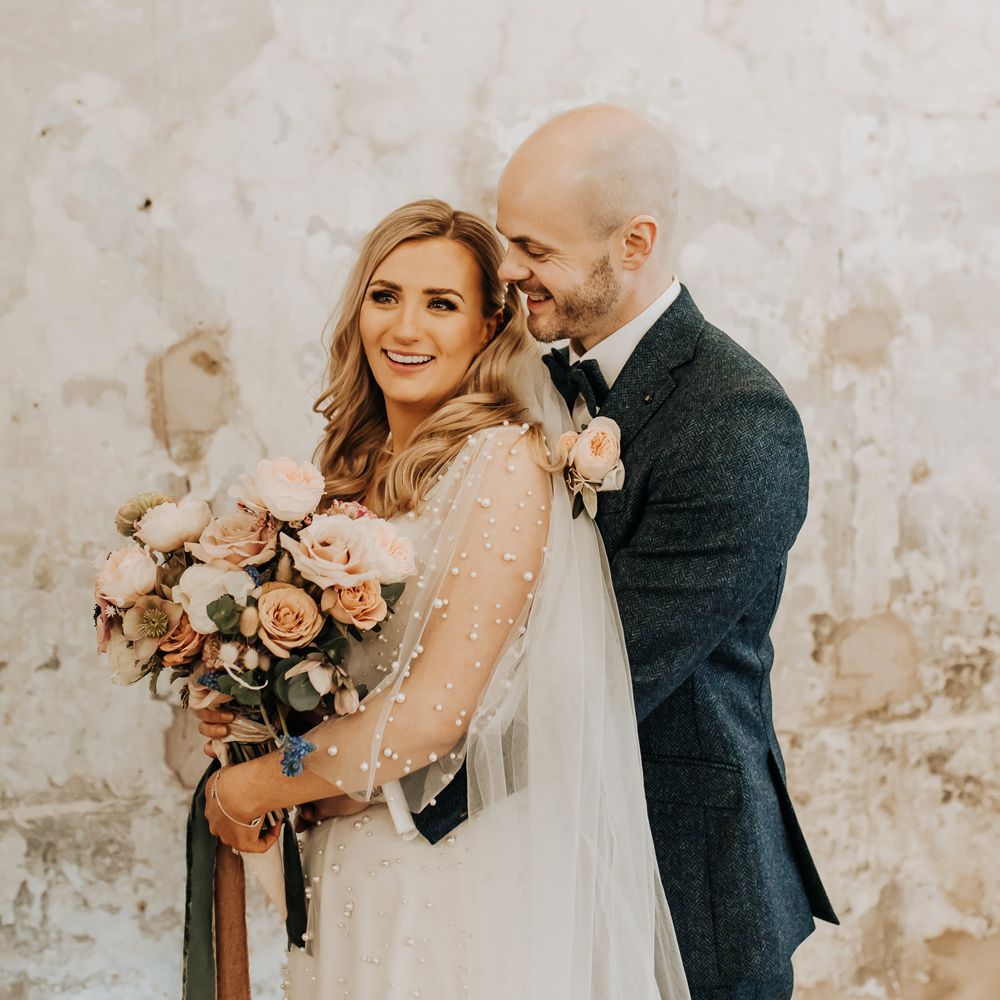 Bride in pearl wedding dress with mesh sleeves stands holding blush pink bridal bouquet with roses and anemones as groom in navy tweed suit hugs her