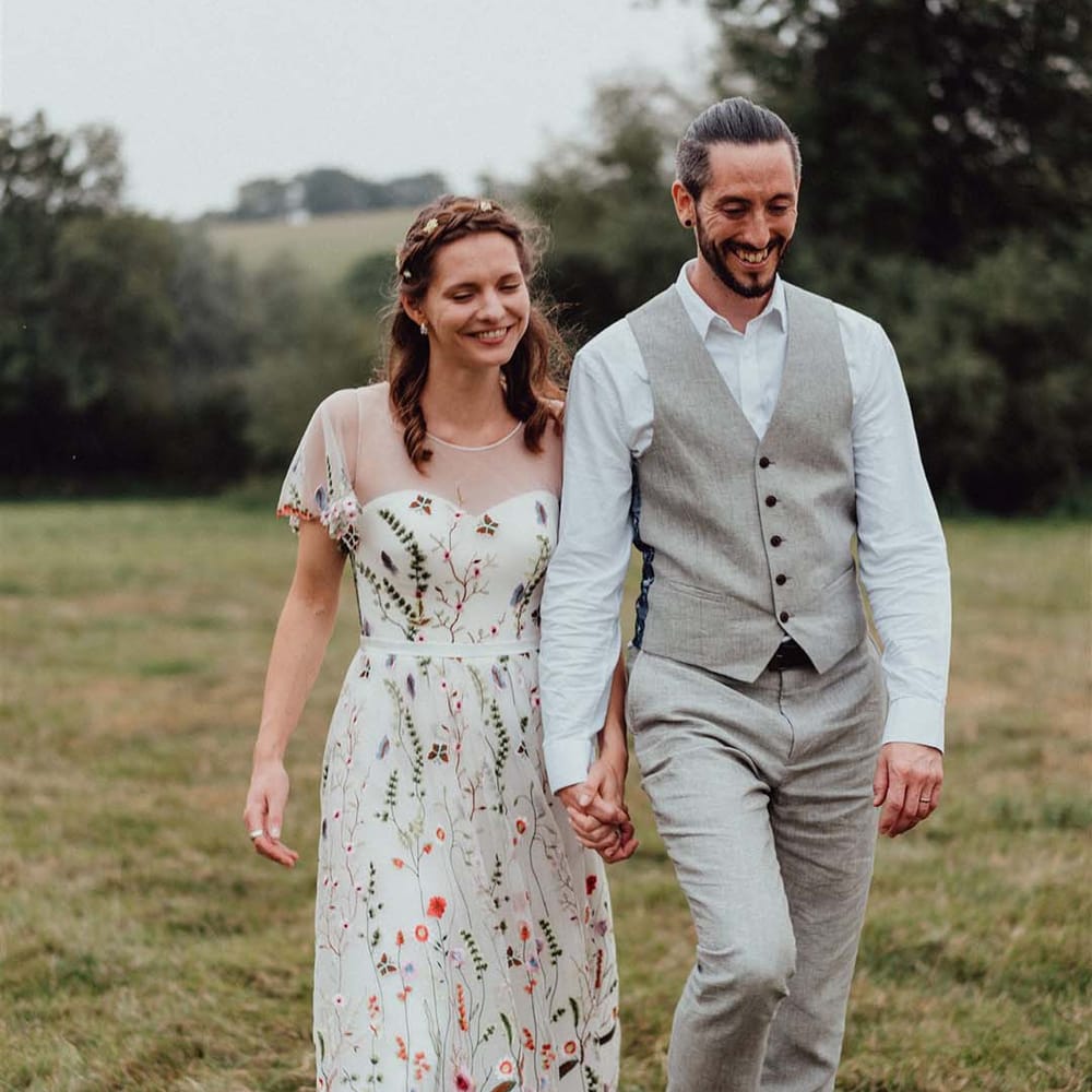 Bride in floral embroidery wedding dress with braided hair and flower accessories with groom in white shirt and grey trousers and waistcoat