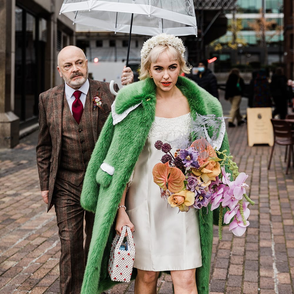 Father of the bride in checkered three piece suit holds an umbrella over the bride as she walks in her platform block heels and green fur coat