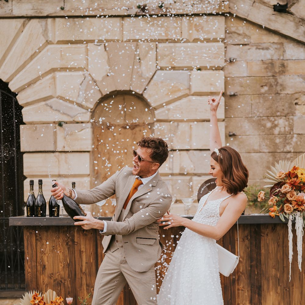 Couple in tartan suit and sleeveless lace wedding dress popping champagne with white confetti