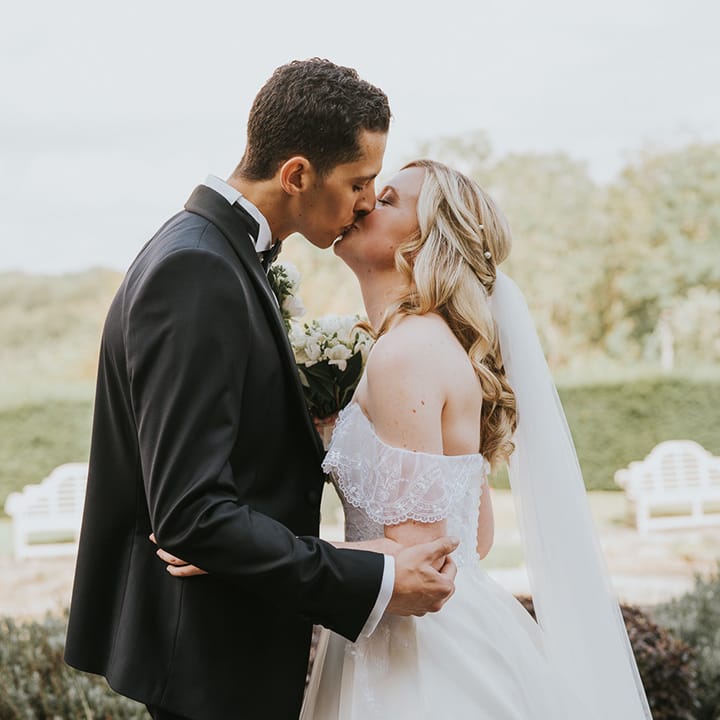 Bride in off the shoulder wedding dress with pearl hair accessory kisses groom in black tie